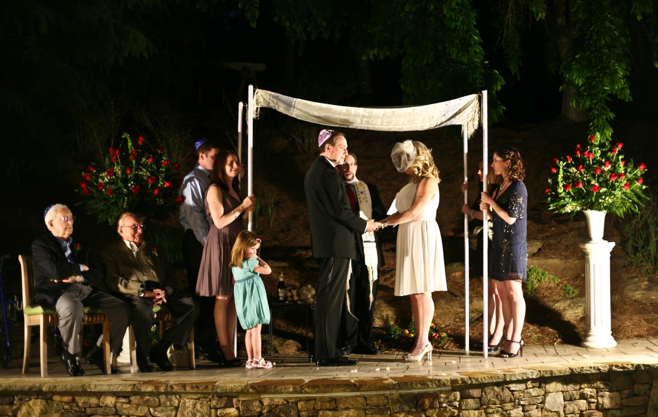 A couple exchanging vows beneath a chuppah at an evening wedding